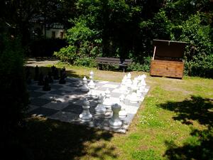 Bild vergrößern: Ansicht auf die Freischachanlage im Grünen mit zirka 60 cm hohen Schachfiguren (hier weiße Schachgfiguren im Vordergrund). Im Hintergund eine Bank zum Verweilen